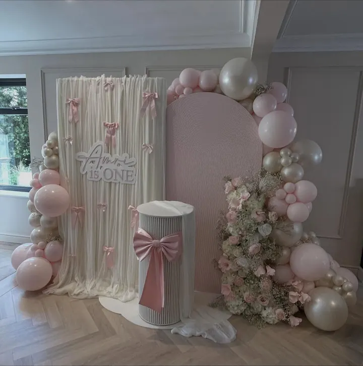 Cream drape and blush arch with pink bows, pearl balloons, florals and a fluted plinth for a first birthday