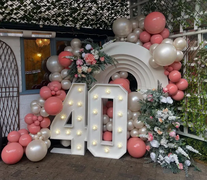 Light-up 40 numbers in front of a white rainbow arch with coral and pearl balloons and faux florals
