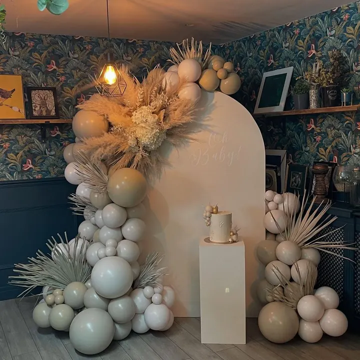 Neutral arch backdrop with sage and cream balloons, pampas and a cake on a plinth