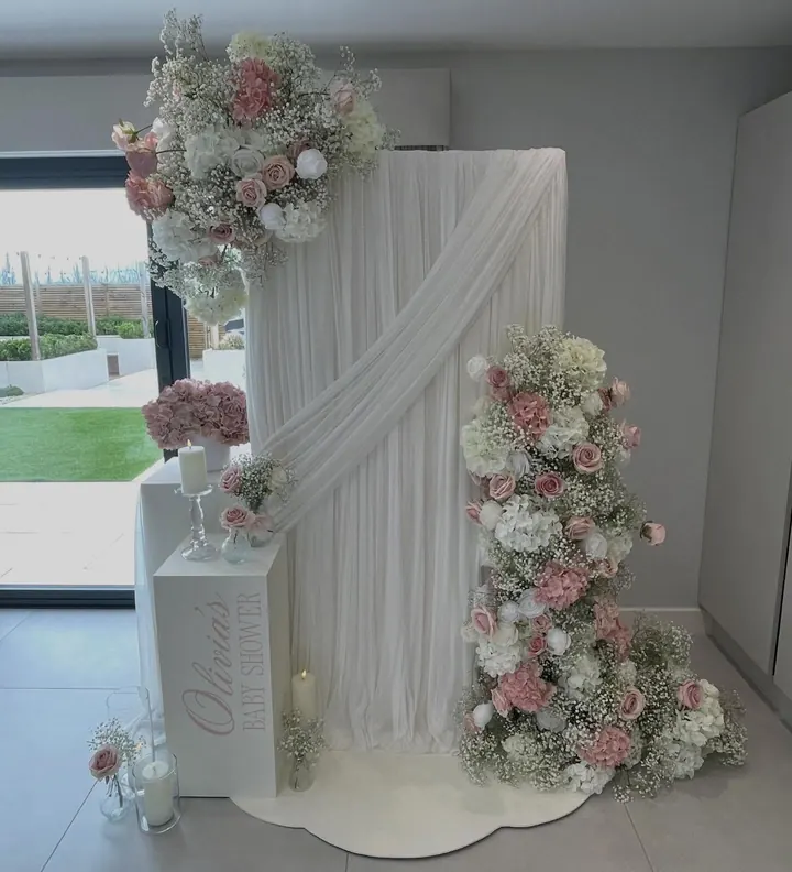 White drape backdrop with pink and white florals and a personalised baby shower plinth
